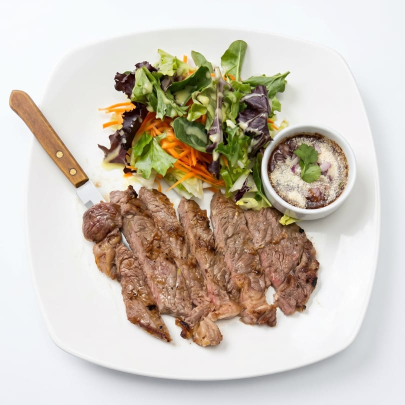 Sliced steak with a side salad, a small bowl of beans, and a steak knife on a white plate.