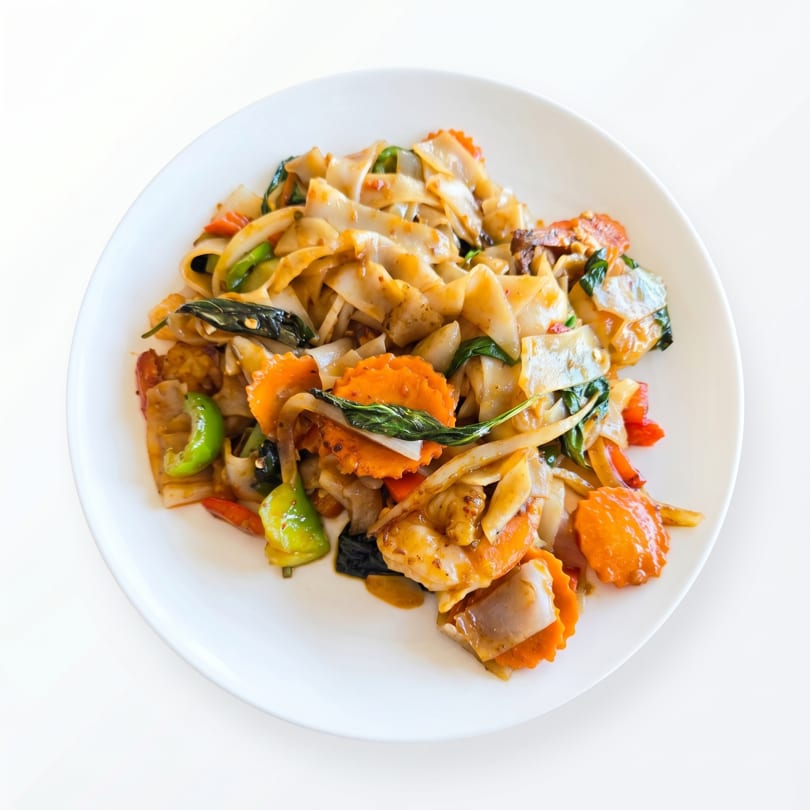 A white plate holds a serving of Pad See Ew with wide noodles, carrots, and green vegetables.